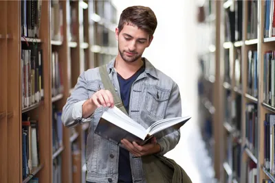 Seorang pria sedang beridir di lorong rak buku sambil membaca buku.