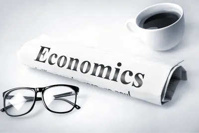 Kertas bertuliskan "ECONOMICS" dengan kacamata dan secangkir kopi di sekelilingnya.